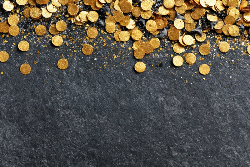 Gold metallic confetti and glitter scattered across a dark stone surface with copyspace below