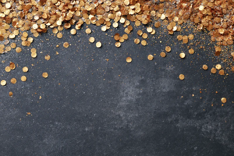 Gold metallic confetti and glitter scattered across dark textured surface with copyspace below