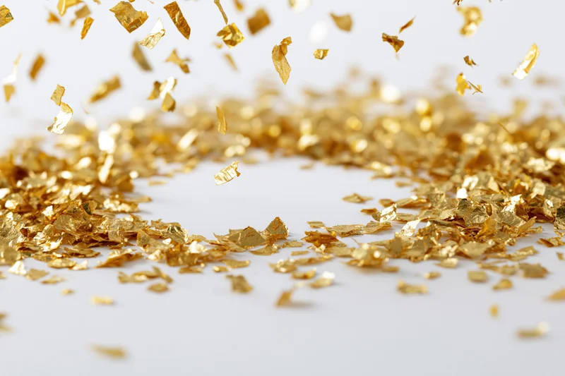 Gold metallic confetti pieces falling and scattering midair on a white surface