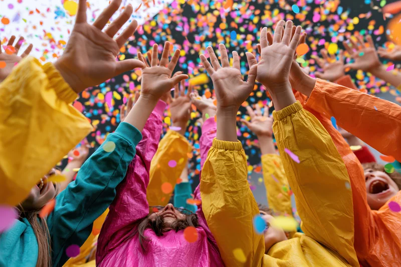 Group of children and adults raising hands upward in colorful confetti at a festive outdoor celebration
