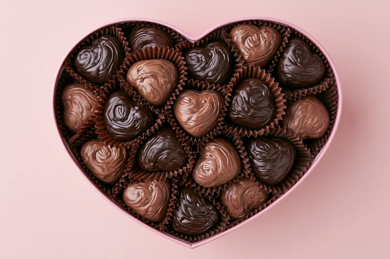 Heart-shaped box filled with assorted chocolate candies on pink background
