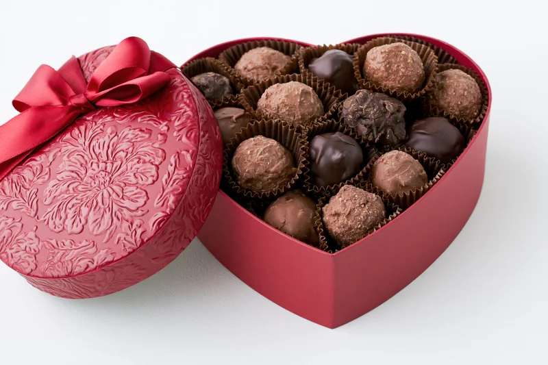 Heart-shaped red gift box filled with assorted chocolate truffles, lid open with ribbon, on white background