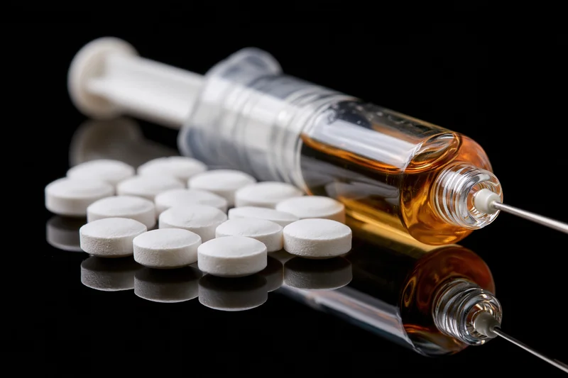 Syringe filled with amber liquid lying beside scattered white pills on reflective black surface