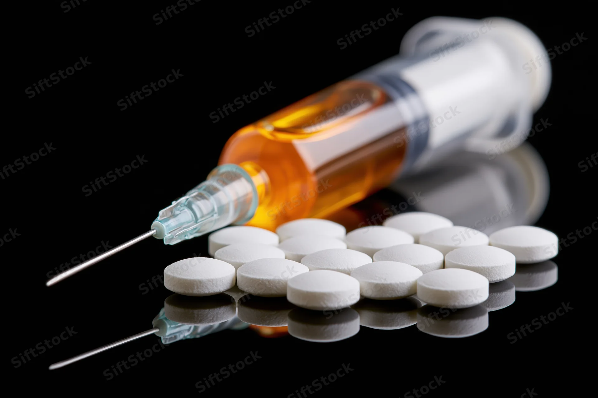 Syringe filled with amber liquid lying beside scattered white tablets on reflective black surface