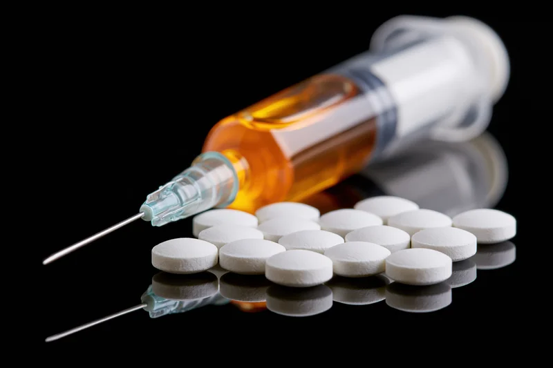 Syringe filled with amber liquid lying beside scattered white tablets on reflective black surface
