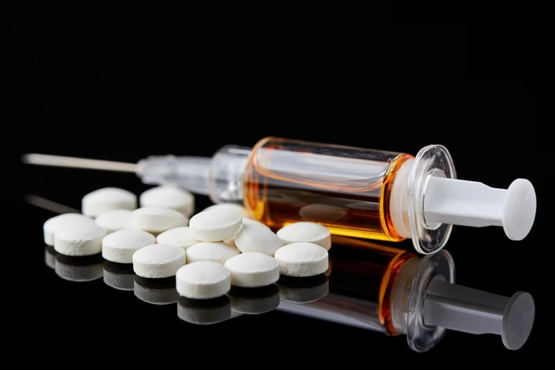 Medical syringe filled with amber liquid lying beside scattered white pills on reflective black surface