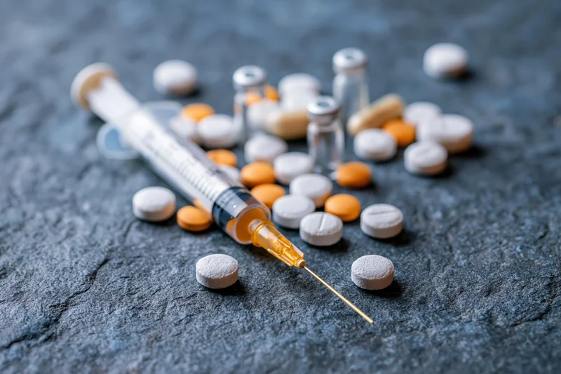 Syringe with gold needle lying among scattered white and orange pills on dark stone surface