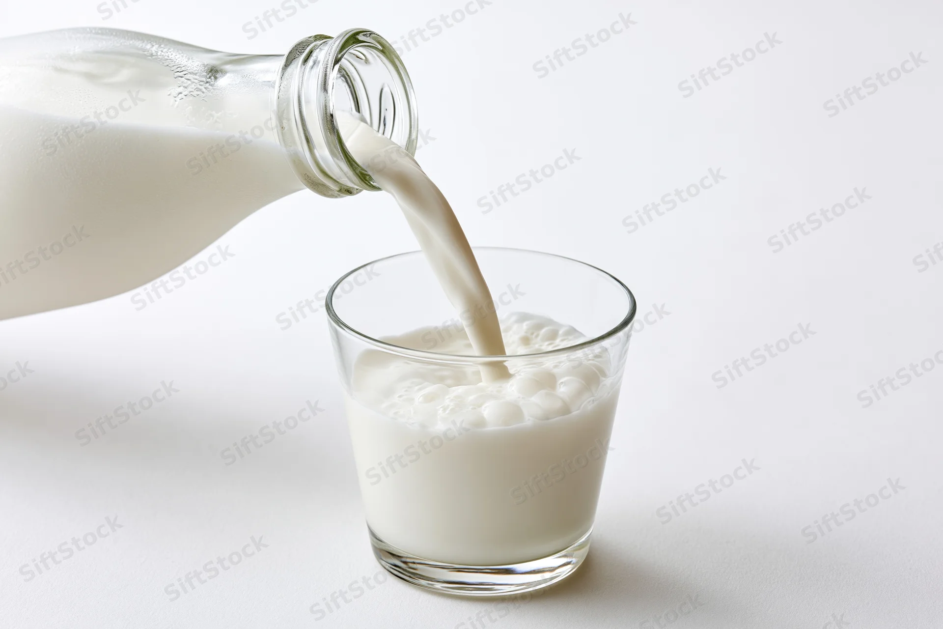 Milk being poured from a clear glass bottle into a small glass on a white surface