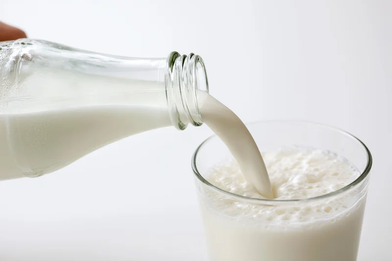Milk being poured from a clear glass bottle into a glass against a white background