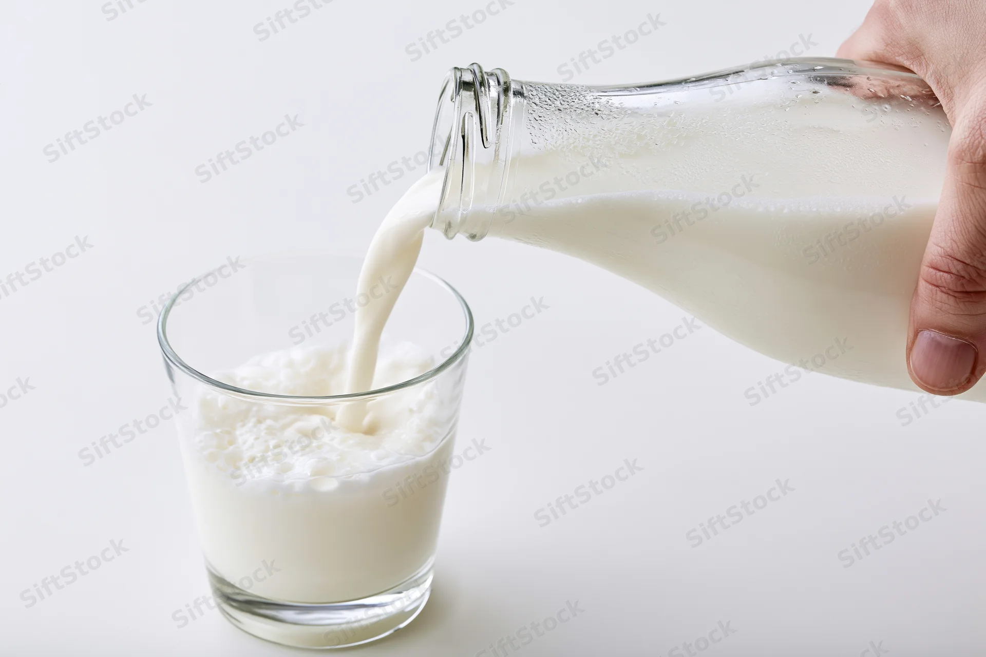 Hand pouring milk from a clear glass bottle into a small glass on a white surface