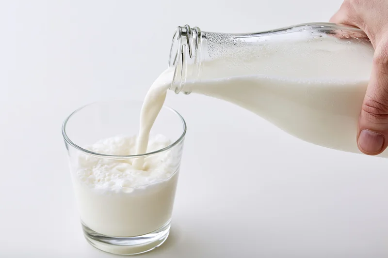 Hand pouring milk from a clear glass bottle into a small glass on a white surface