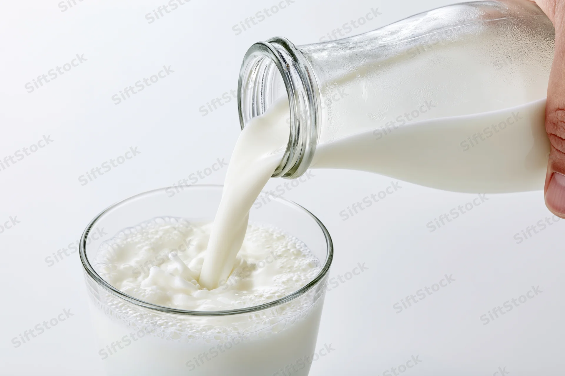 Hand pouring milk from a clear glass bottle into a glass on a white background