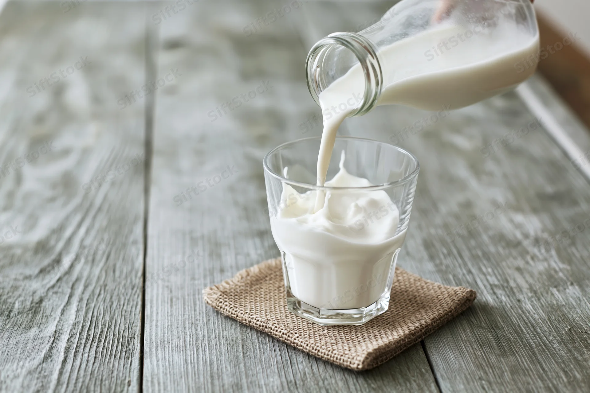 Milk being poured from a glass bottle into a clear drinking glass on a wooden table