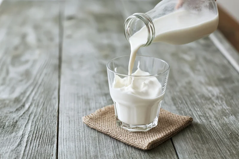 Milk being poured from a glass bottle into a clear drinking glass on a wooden table