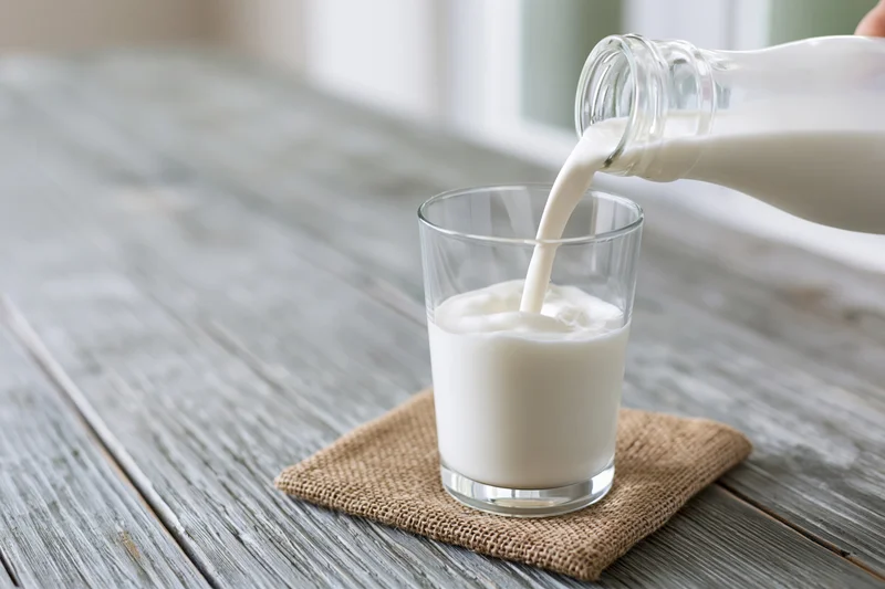 Milk being poured from a glass bottle into a clear glass on a wooden table