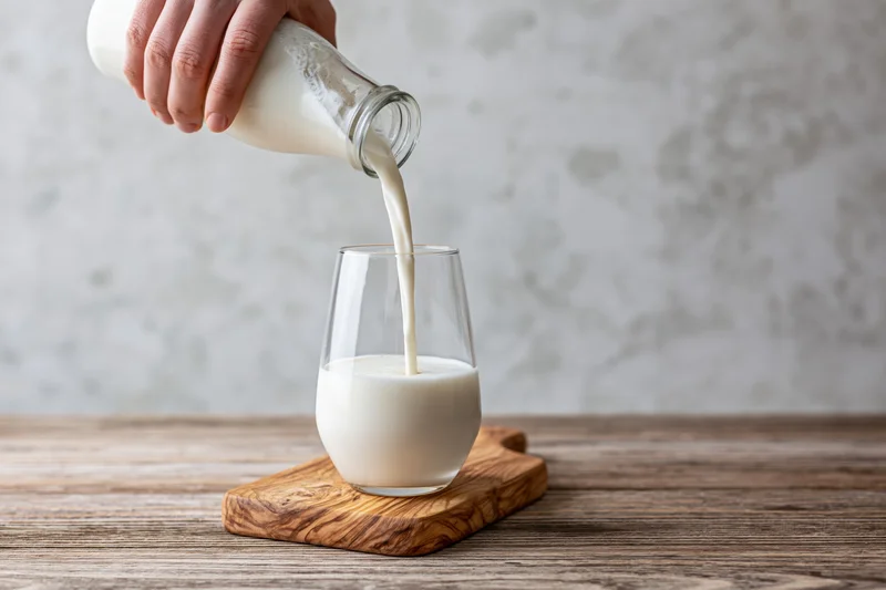 Hand pouring milk from a glass bottle into a stemless glass on a wooden cutting board