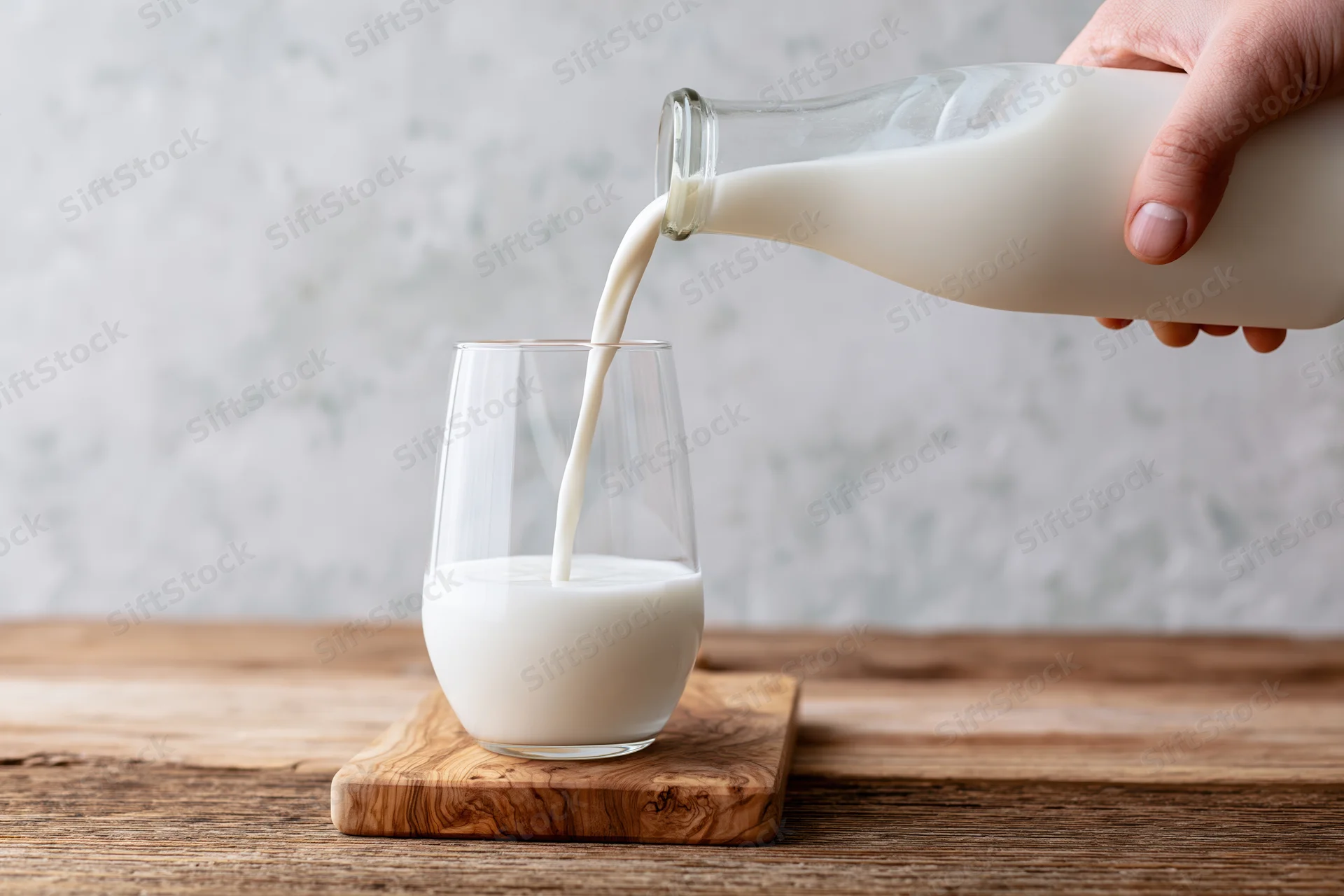 Hand pouring milk from a glass bottle into a clear stemless glass on a wooden cutting board