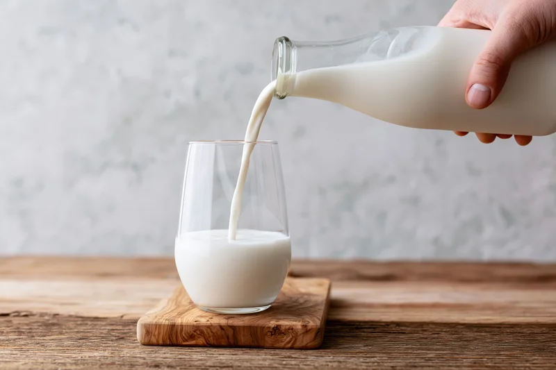 Hand pouring milk from a glass bottle into a clear stemless glass on a wooden cutting board