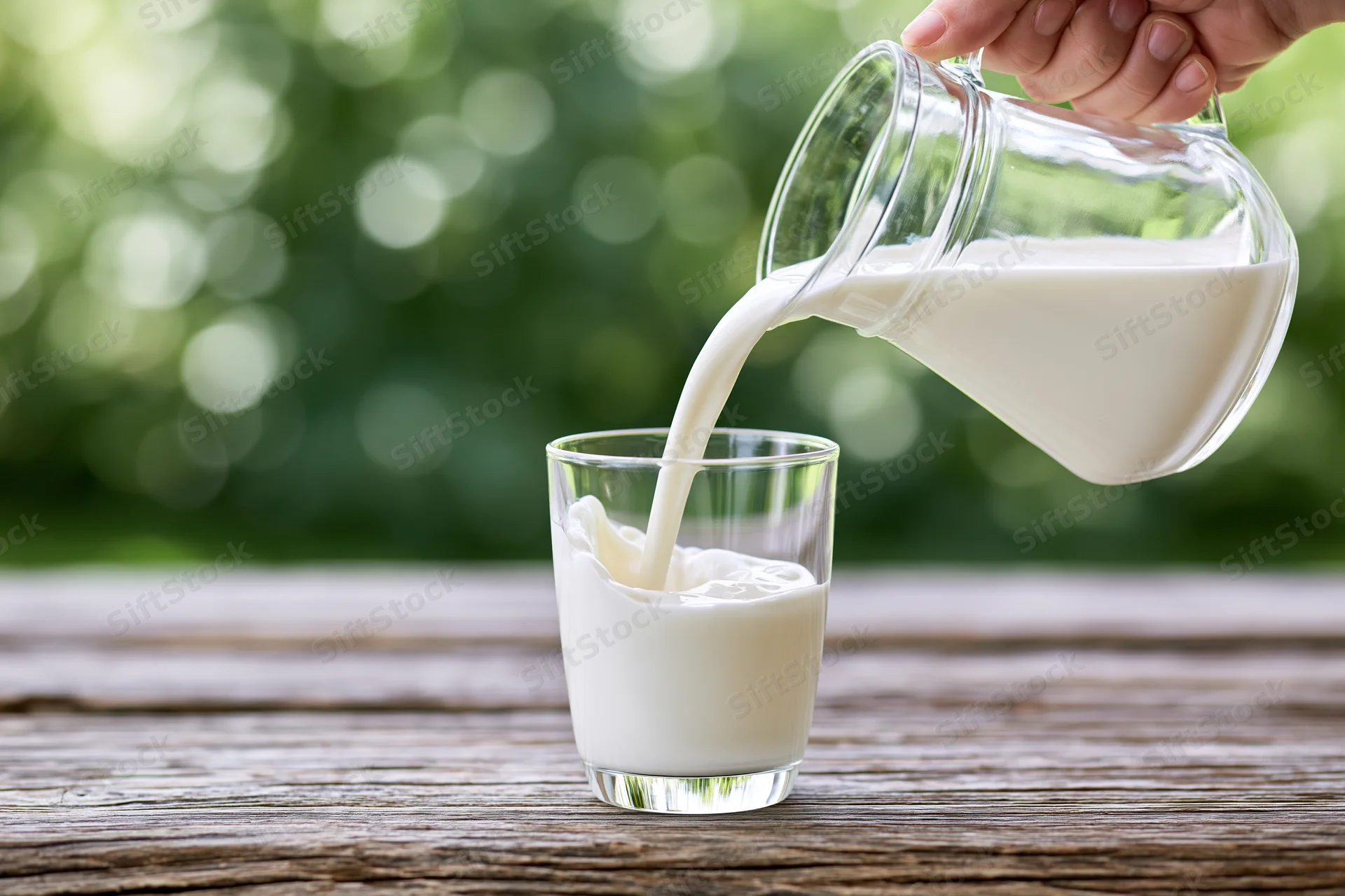 Hand pouring milk from a glass pitcher into a clear glass on a wooden table outdoors