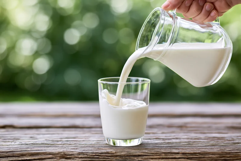Hand pouring milk from a glass pitcher into a clear glass on a wooden table outdoors