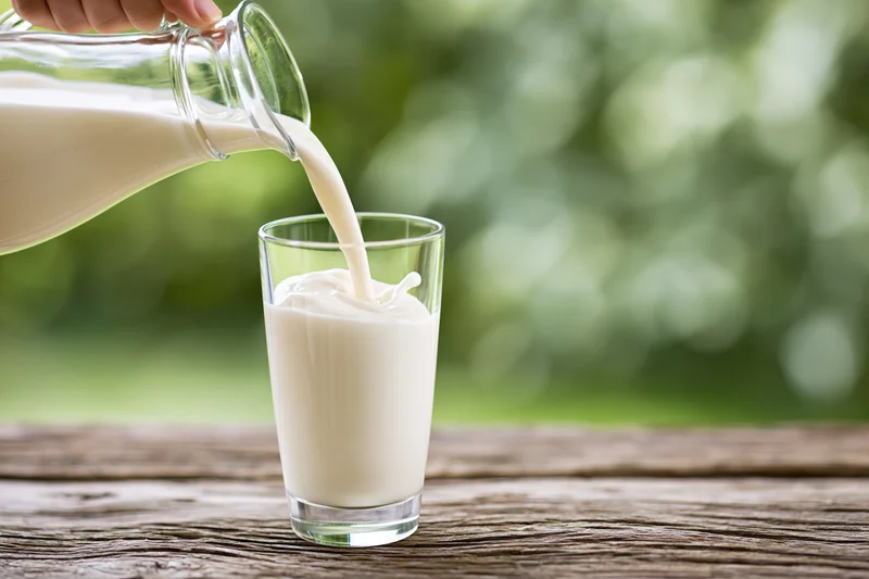 Milk being poured from a glass pitcher into a clear drinking glass on a wooden table outdoors