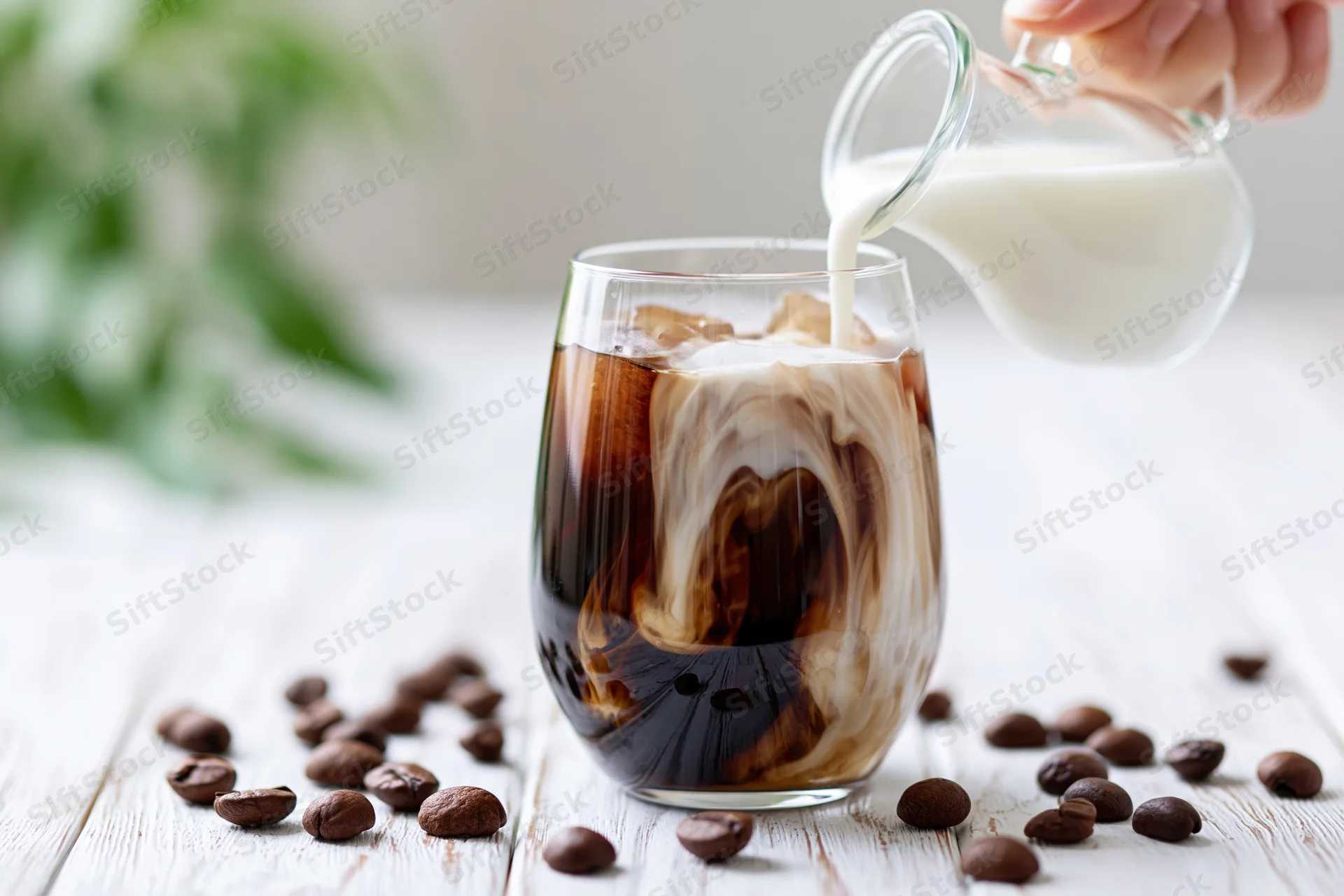 Milk being poured into a glass of iced coffee on a white wooden table surrounded by coffee beans