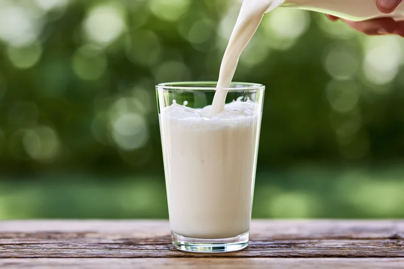 Milk being poured into a clear glass tumbler on a wooden table outdoors