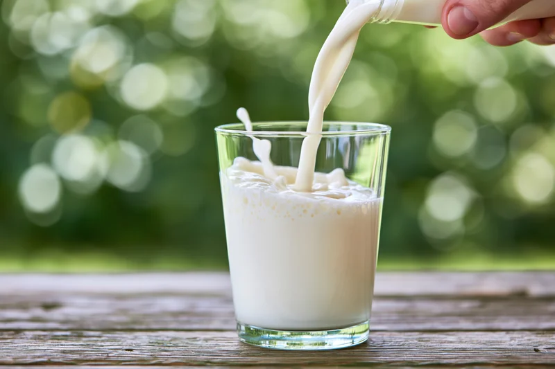 Milk being poured into a clear glass tumbler on a wooden table outdoors