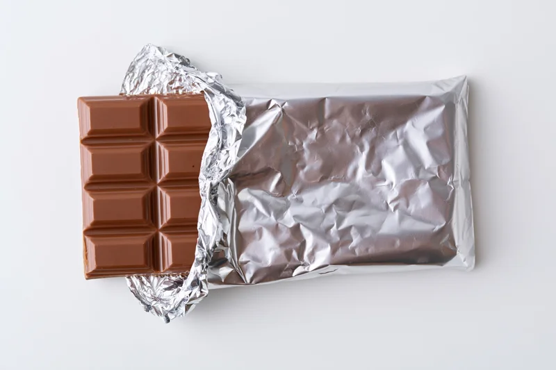 Partially unwrapped milk chocolate bar on white background with crinkled silver foil wrapper