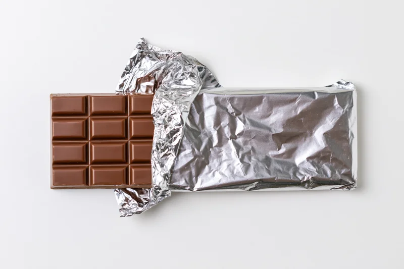 Partially unwrapped milk chocolate bar on white background with crinkled silver foil wrapper