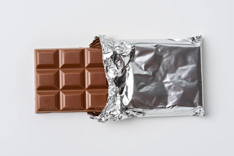 Partially unwrapped milk chocolate bar with crinkled silver foil wrapper on white background
