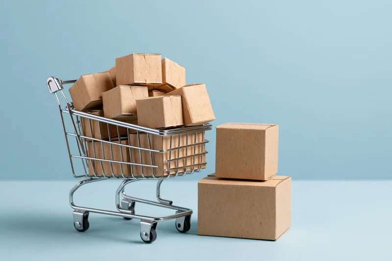Miniature metal shopping cart filled with small cardboard boxes on light blue surface