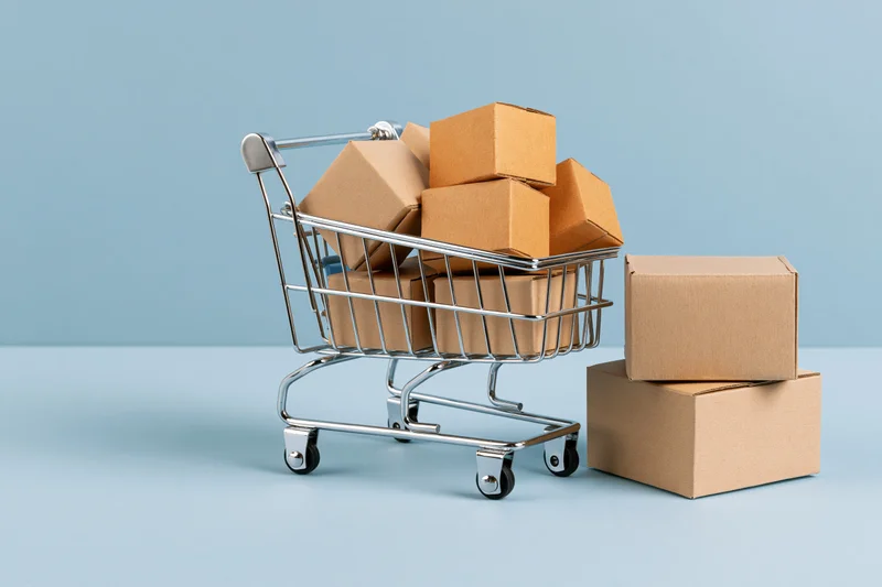 Miniature metal shopping cart filled with small cardboard boxes on a light blue surface