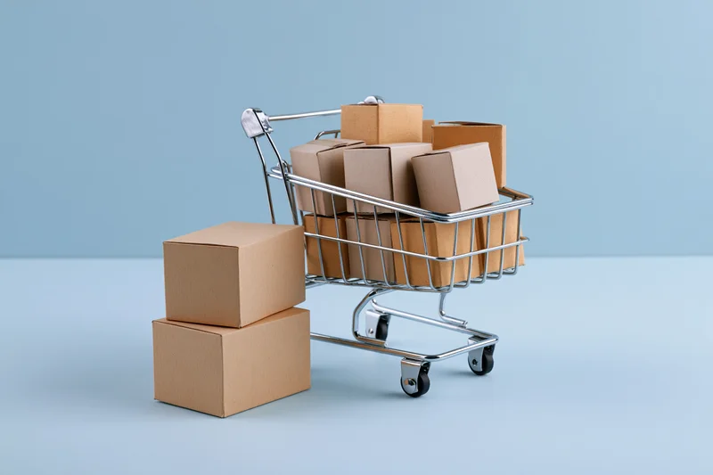 Miniature metal shopping cart filled with small cardboard boxes on a light blue surface