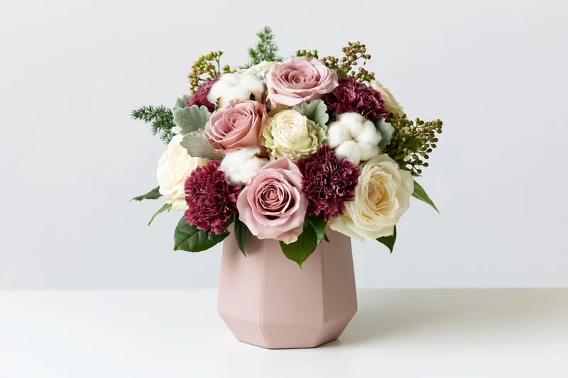 Mixed flower bouquet with roses, carnations, cotton and greenery in a pale pink geometric vase on a white surface