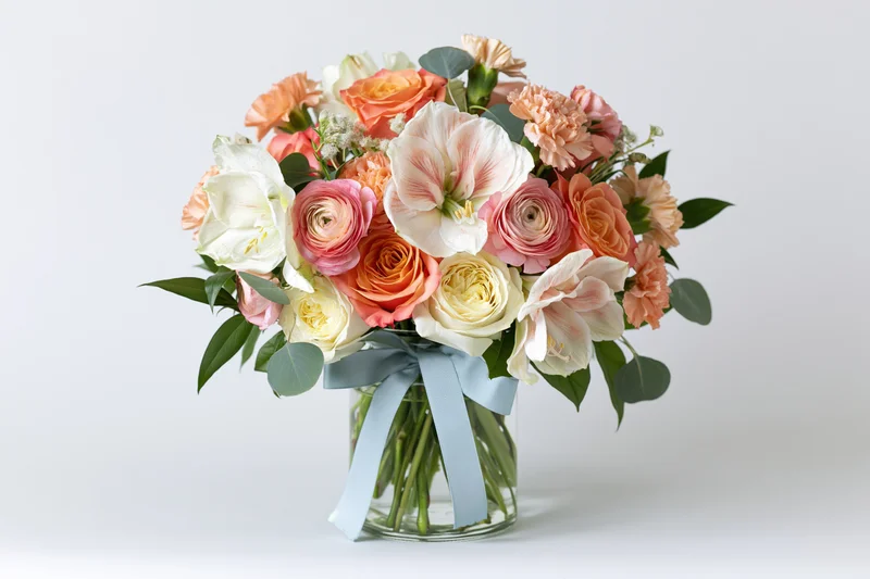 Mixed spring flower bouquet in a clear glass vase with blue ribbon centered on white background