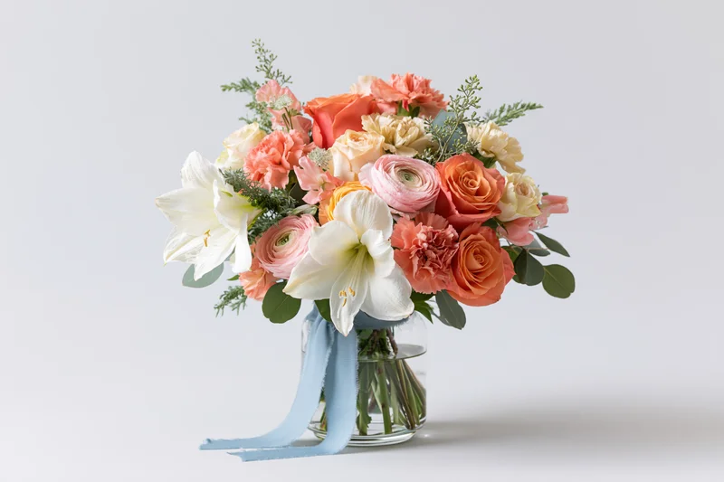 Mixed spring flower bouquet in a clear glass vase with blue ribbon, centered on white background