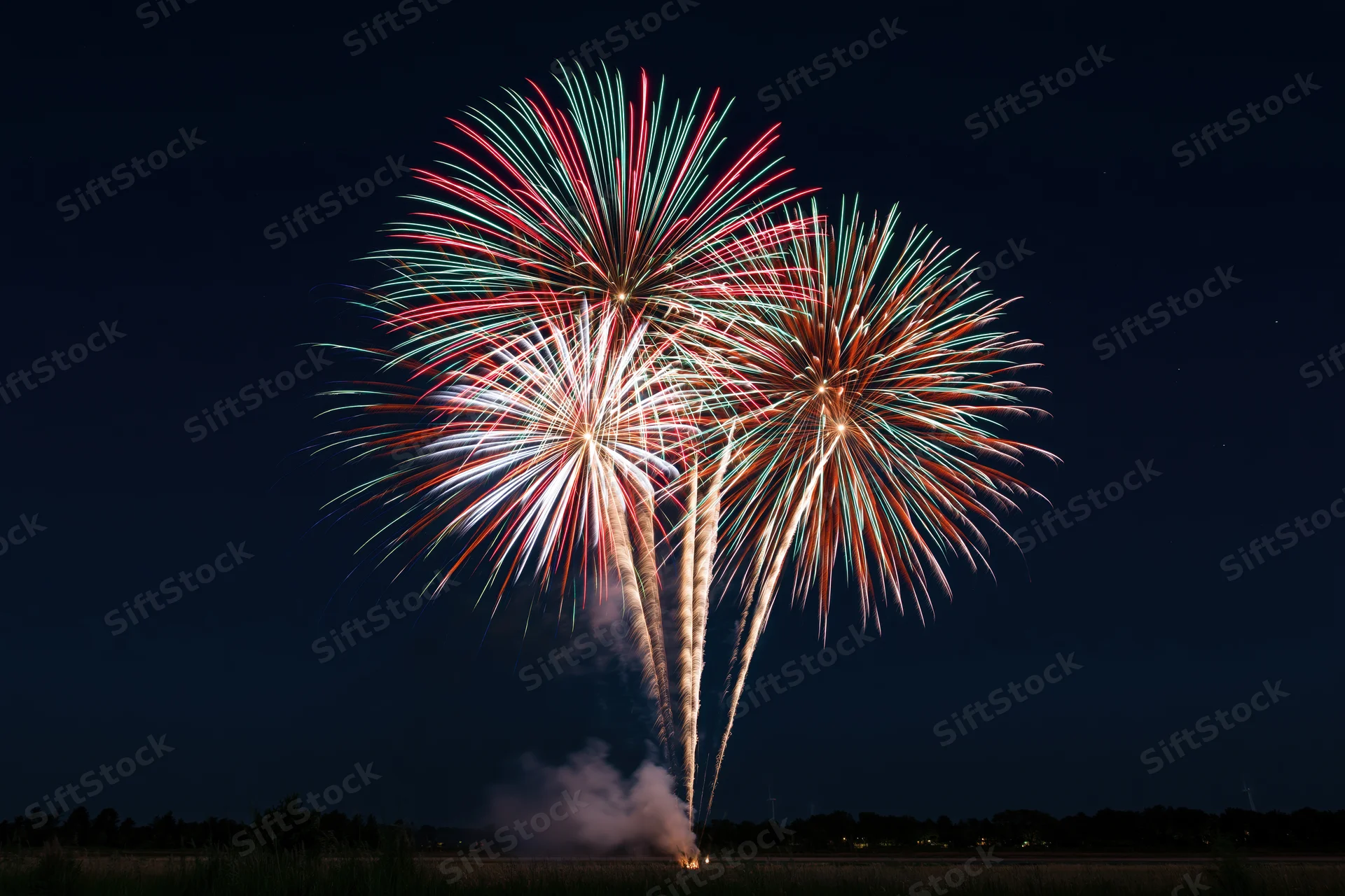 Multiple fireworks bursting simultaneously in the dark night sky over an open field