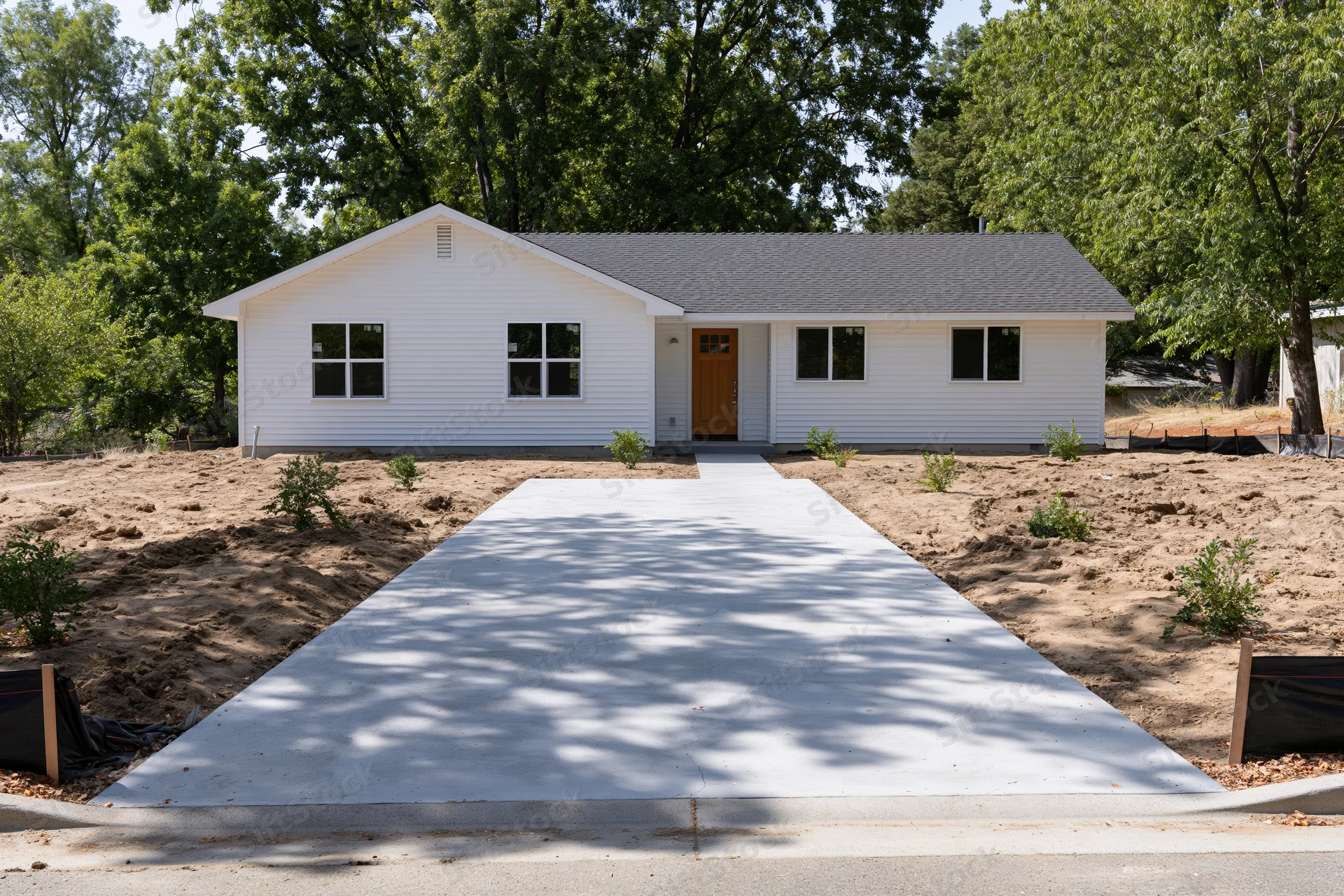 Newly poured concrete driveway leading to a white single-story house with front door and windows, surrounded by trees and bare soil in suburban yard
