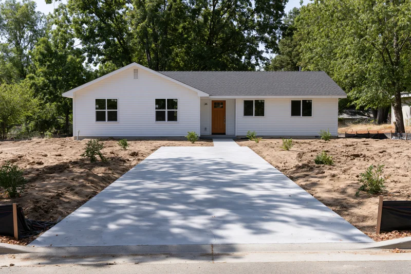 Newly poured concrete driveway leading to a white single-story house with front door and windows, surrounded by trees and bare soil in suburban yard