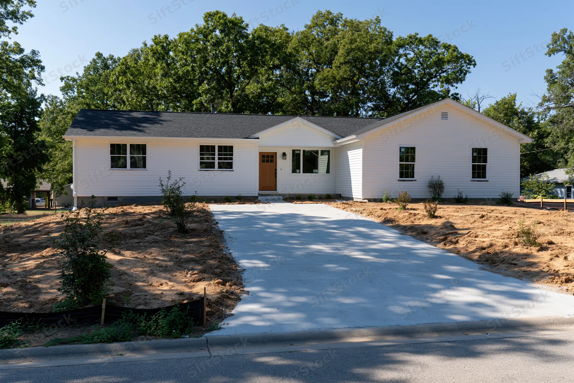 White single-story house with newly poured concrete driveway leading to front entrance, surrounded by trees and dirt yard under clear blue sky