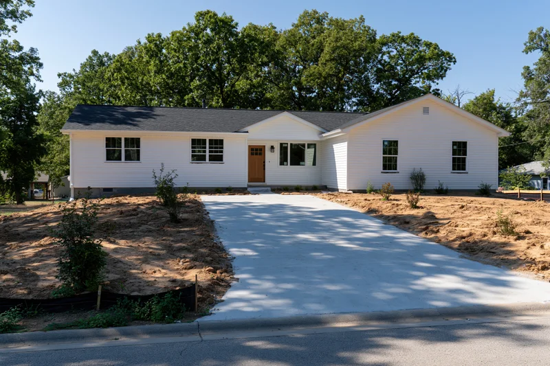 White single-story house with newly poured concrete driveway leading to front entrance, surrounded by trees and dirt yard under clear blue sky