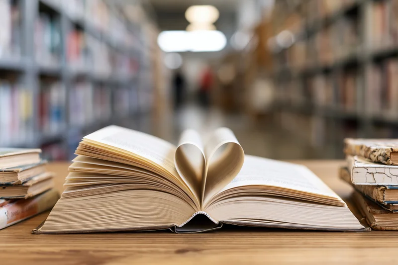 Open hardcover book with pages folded into heart shape on wooden table in library aisle