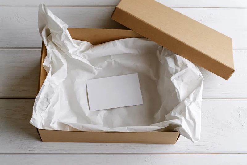 Open kraft cardboard gift box lined with white tissue paper on wooden surface with blank card inside