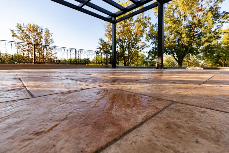 Wet stamped concrete patio under metal pergola with trees in background