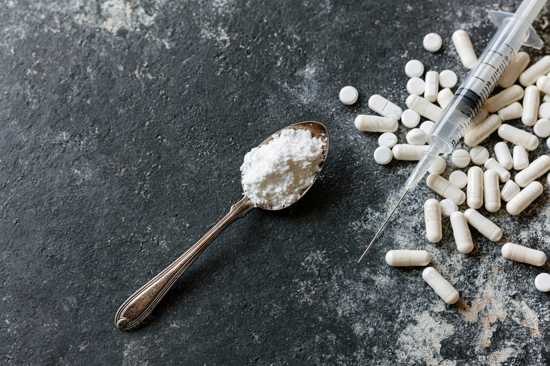 Tarnished spoon holding white powder with syringe and scattered pills on dark textured surface
