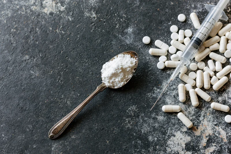 Tarnished spoon holding white powder with syringe and scattered pills on dark textured surface