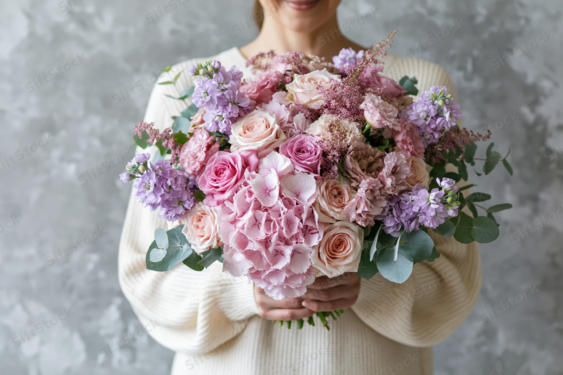 Woman holding a large lush bouquet of pink and purple flowers with both hands against a textured gray background