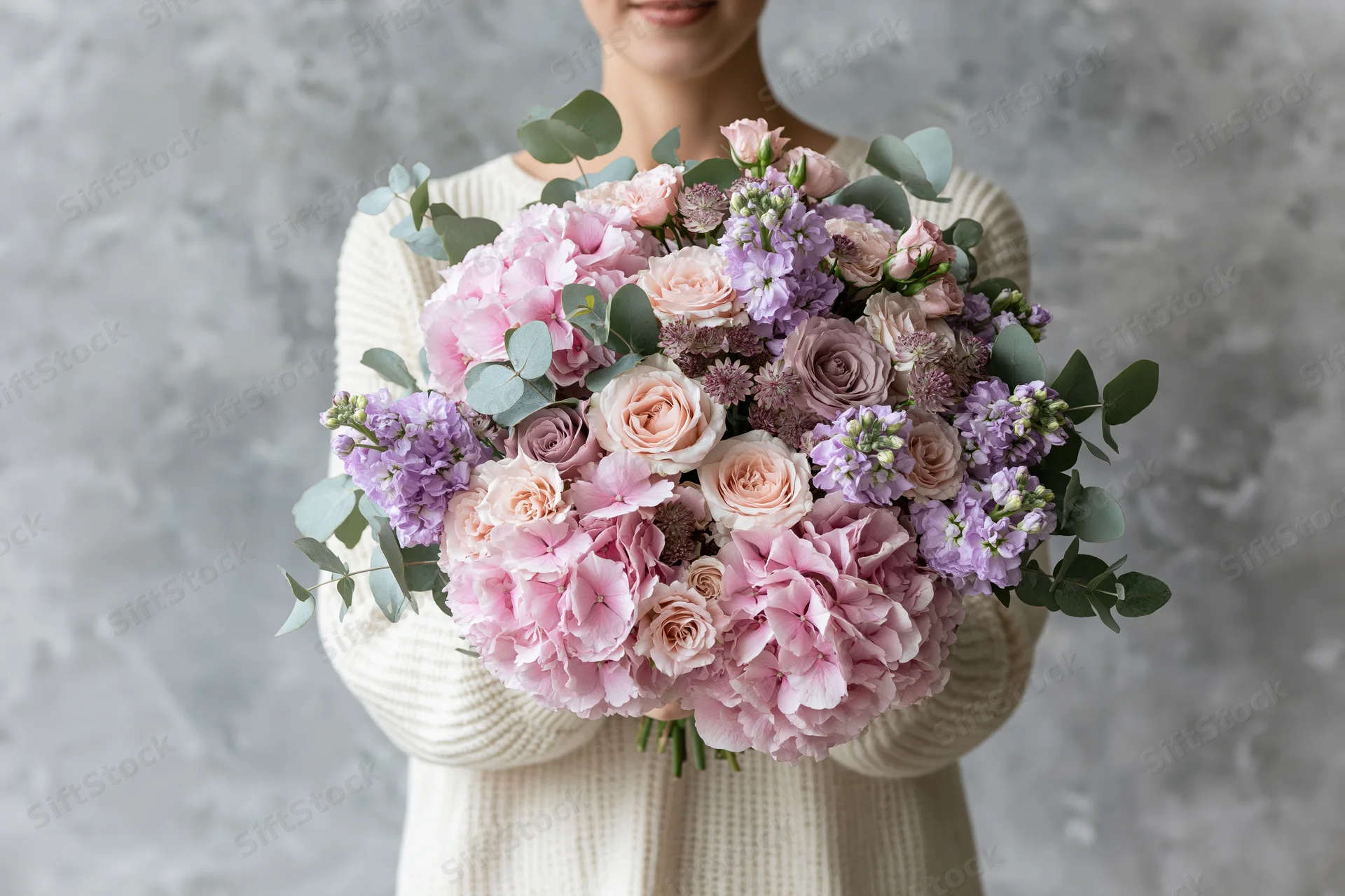 Woman holding large lush bouquet of pink and purple flowers with greenery, smiling, partial face visible, cream sweater, gray textured background