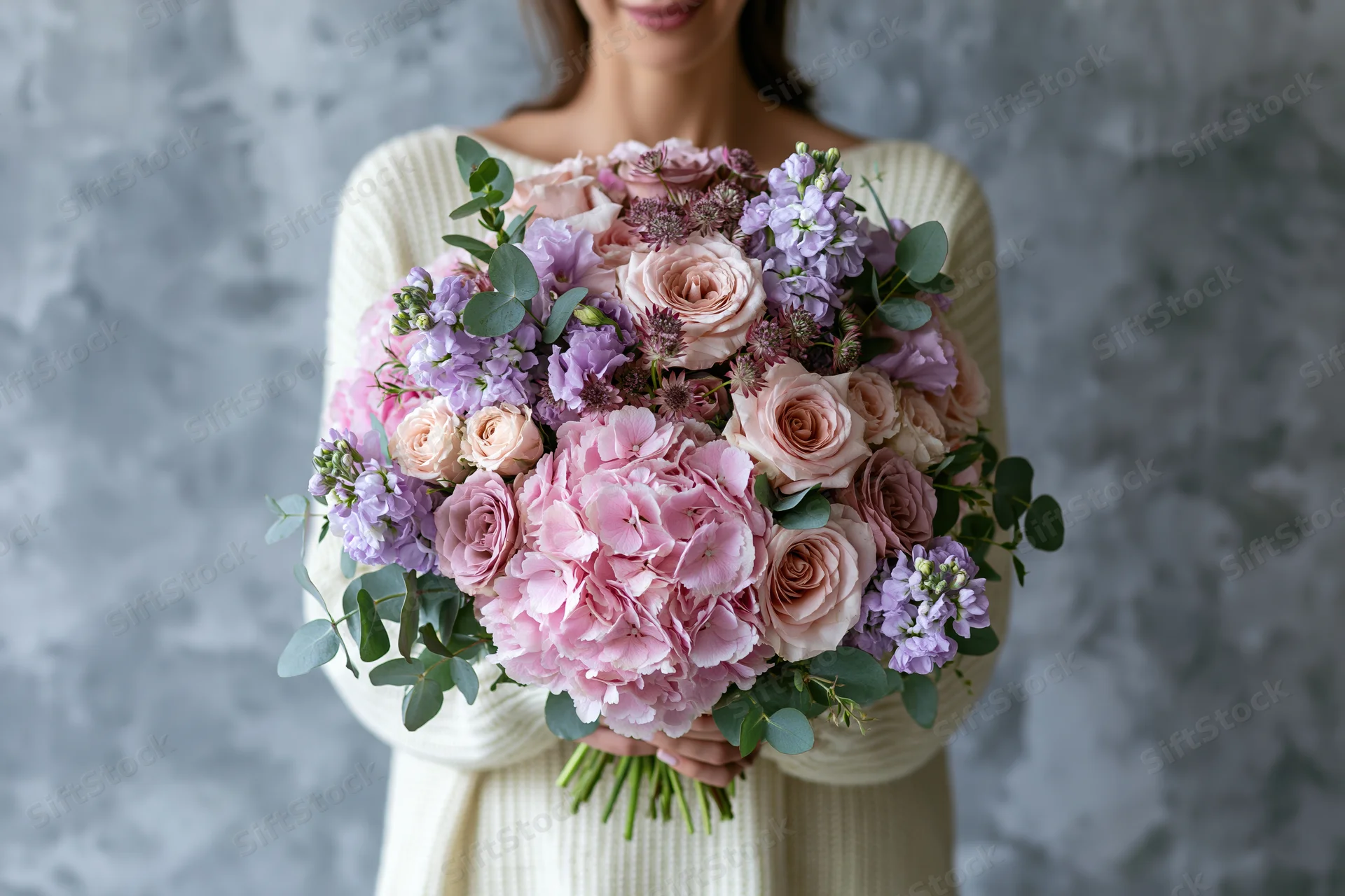 Woman holding a large lush bouquet of pink roses, hydrangea, lavender and eucalyptus against a textured gray background
